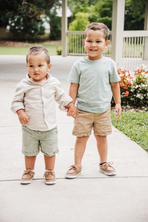 Two small children holding hands while standing on a concrete walkway near flowers.
