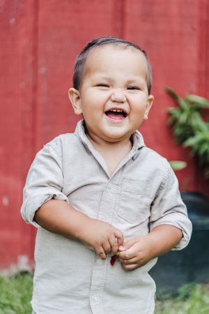 A joyful toddler laughing outdoors against a red barn wall wearing a light colored shirt.