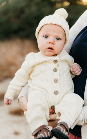 Baby in cream knit outfit with pom beanie and brown shoes looking at camera.