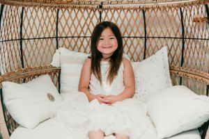 Young person in white dress sitting on wicker chair with white pillows.