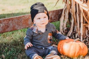 Small child wearing knit hat sits outdoors next to large orange pumpkin on autumn day.