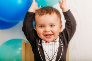 Baby in tuxedo outfit raising hands with blue and teal balloons in background.