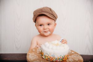 Baby wearing newsboy cap sitting with decorated birthday cake during cake smash photo session.