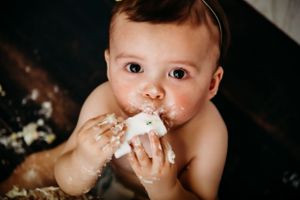 Close-up of messy cake smash photography with white frosting covering a baby's mouth and hands.
