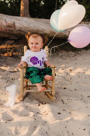 Toddler in mermaid themed outfit sits in wooden rocking chair with pastel balloons.