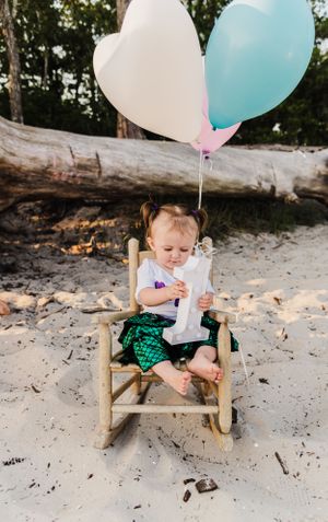 Small child in mermaid outfit sitting in wooden rocking chair with heart balloons.