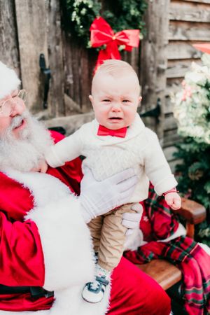 Baby in white sweater crying during holiday photo session with Santa.