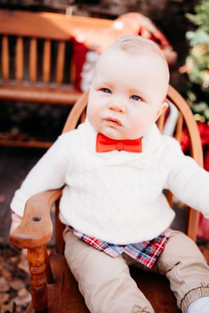 Baby in formal white sweater and plaid shorts with red bowtie sits in wooden rocking chair.
