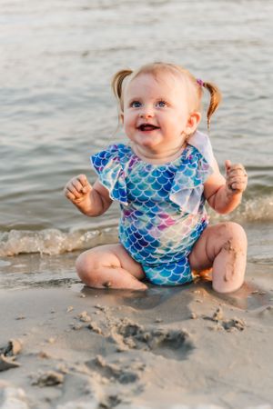 Baby in mermaid swimsuit sitting and playing in wet sand on beach.