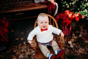 Smiling baby in white outfit with red bowtie sits in wooden chair surrounded by holiday decor.