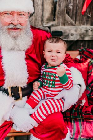 Baby in red and white striped holiday pajamas sits on traditional figure in red suit with white beard for Christmas photo.