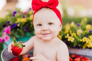 Sweet baby wearing red bow headband holds fresh strawberry while sitting among colorful garden flowers.