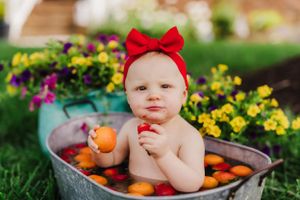 A small figure wearing a red bow sits in a metal tub filled with oranges on green grass near colorful flowers.