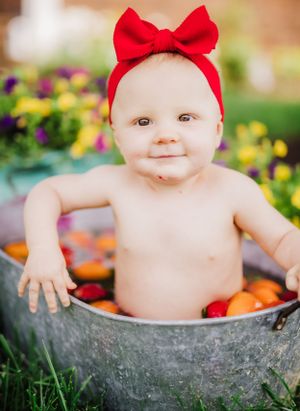 A baby wearing a red bow headband sits in a vintage metal tub surrounded by colorful flowers in a garden setting.