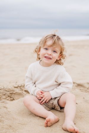 Toddler in white sweater sitting on sandy beach with bright smile.