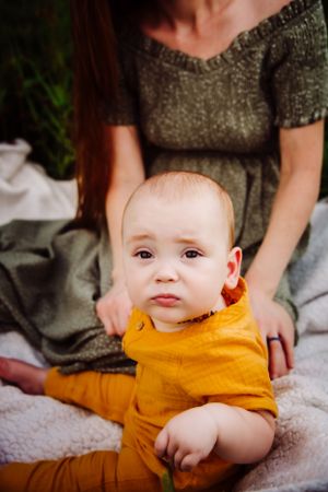 Baby in yellow outfit sitting on blanket with caregiver.
