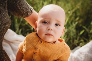 Baby in mustard yellow button-up shirt sitting outdoors on white blanket.