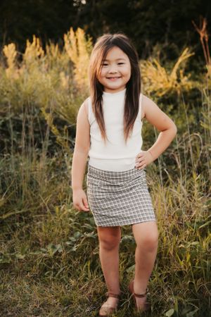 Young person in white sleeveless top and plaid skirt poses confidently outdoors.