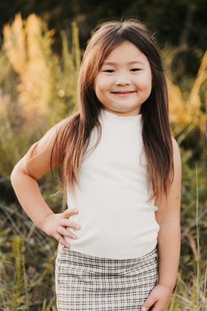 A child wearing a white sleeveless top and plaid skirt poses outdoors against golden grass in warm sunlight.