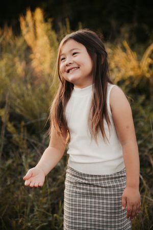 A child in a white top and plaid skirt smiles joyfully while standing in a field at golden hour.