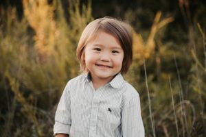 A young child in a striped button-down shirt smiles brightly against a backdrop of golden wild grass.