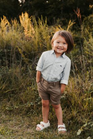 A child in a striped shirt and brown shorts stands outdoors among tall golden grasses at sunset.