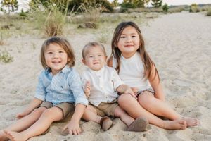 Three children sit together smiling on a sandy beach during a family photo session.