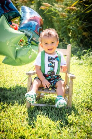 Small child in green suspenders and printed shorts sits in wooden chair outdoors with balloons.