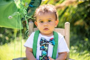 Child wearing white shirt with green suspenders sits in wooden chair in sunny garden setting.