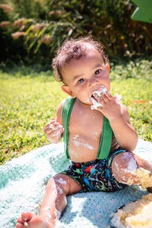 Young child in green suspenders enjoys messy cake smash photoshoot outdoors.