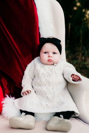 Baby wearing white knit dress and black headband sitting on velvet chair.