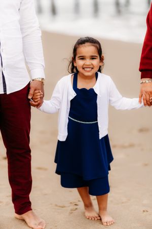 Little girl in navy dress and white cardigan holding hands on beach.