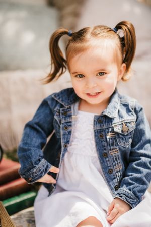 Adorable toddler in denim jacket and pigtails smiles sweetly at camera.