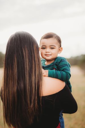 A toddler in a green sweater smiles while being held by someone with long dark hair in an outdoor setting.