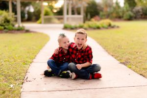 Two children in matching buffalo plaid shirts sitting on concrete pathway.