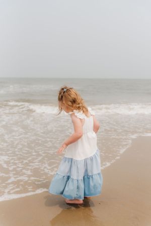Young girl in tiered dress playing at ocean's edge on sandy beach.