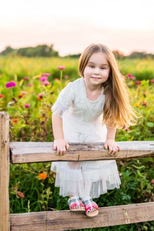 Long-haired figure in white dress leaning on wooden fence surrounded by pink flowers.