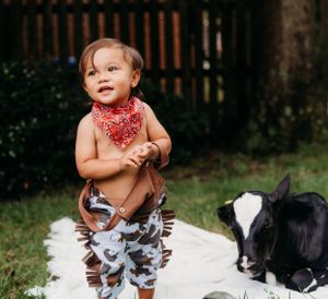 Child in bandana and patterned pants sitting on blanket with black and white goat.