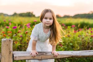 Child in white dress leaning on wooden fence post among pink summer flowers.