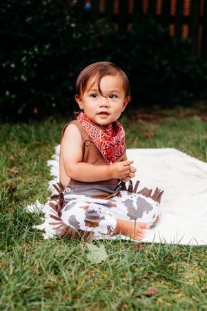 Child wearing red bandana and cow print pants sitting on white blanket outdoors.