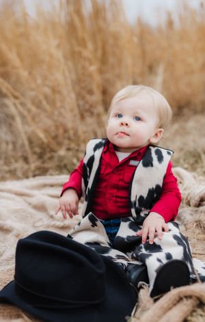 A baby in cow print vest and red shirt sits on blanket surrounded by tall grass.