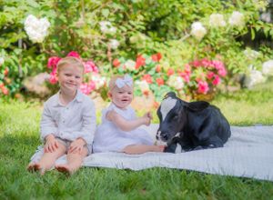 Two children sit with a black and white calf on a white blanket in a flowering garden.