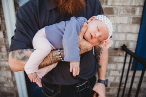 Sleeping baby in blue outfit rests peacefully on tattooed arms.