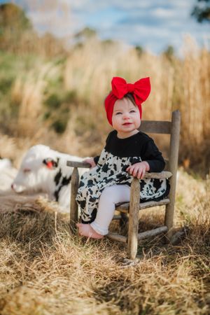 A small child in a black and white dress with a red bow sits in a wooden chair outdoors.