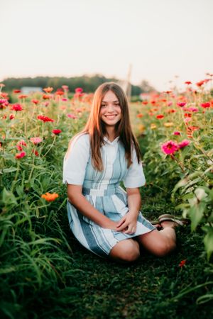 Young girl in plaid dress sitting among blooming red flowers during golden hour.