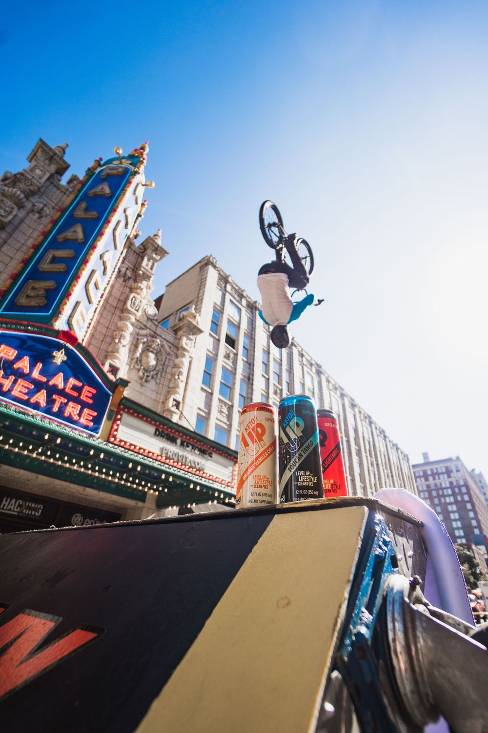 BMX rider performing aerial backflip trick in front of historic Palace Theatre marquee against blue sky.