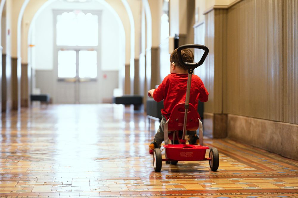 Child riding a red push scooter down a grand hallway with arched ceilings.