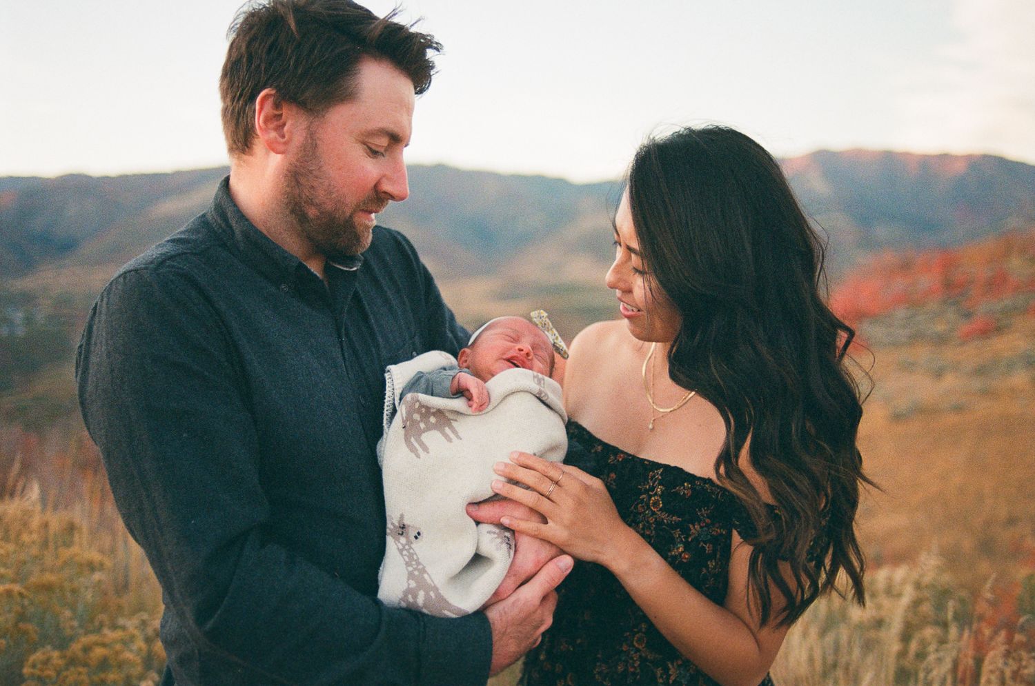 Parents gazing lovingly at newborn baby wrapped in white blanket against autumn backdrop.