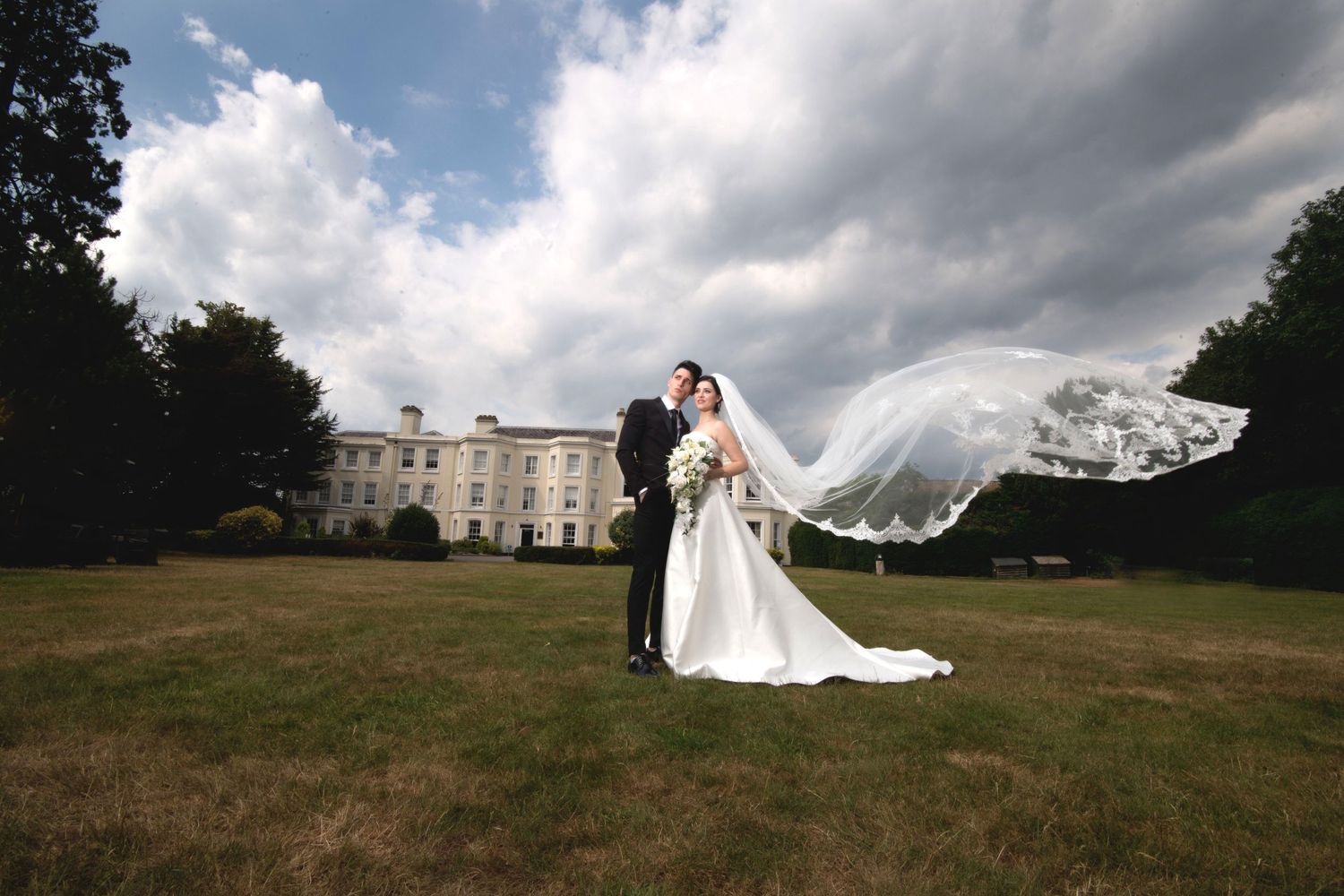 Dramatic wedding photo with bride's veil flowing in wind against stately mansion backdrop.