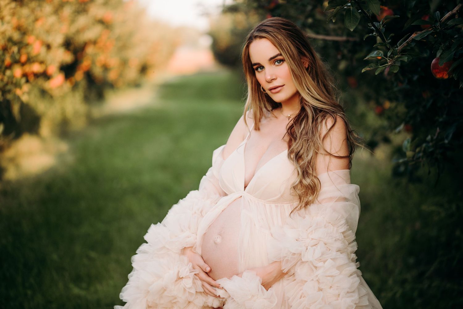 Expectant mother in flowing pink dress posing in an autumn orchard at sunset.